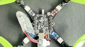 The Crazyflie 2.1 Brushless on the desk, green props, the battery strapped on top, on a black desk mat